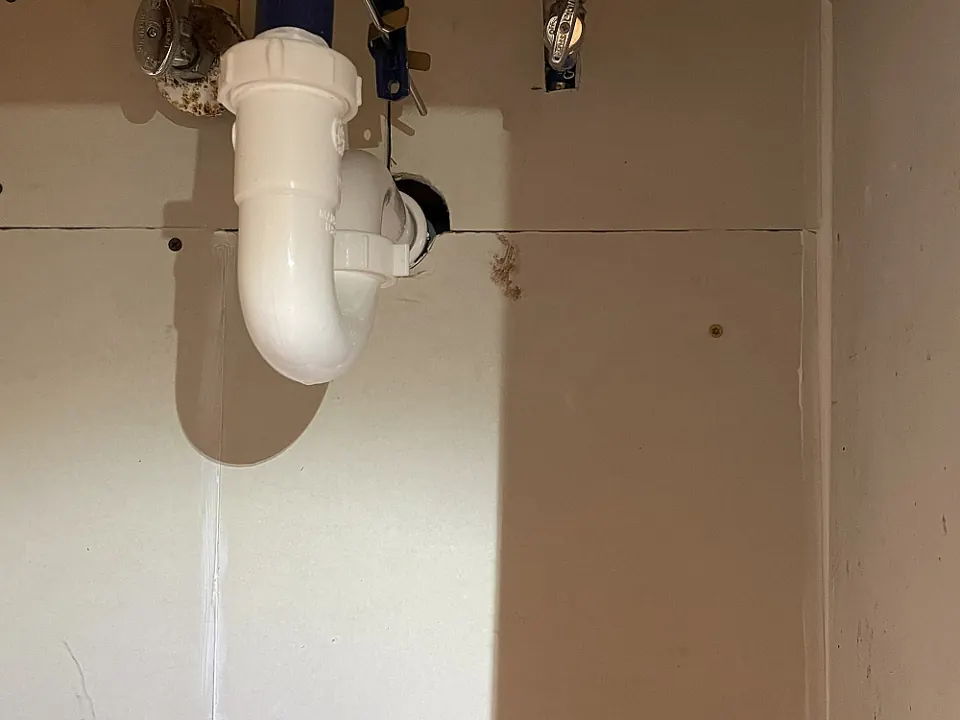 New white PVC drain trap under a bathroom sink, with new sheetrock on the wall behind it